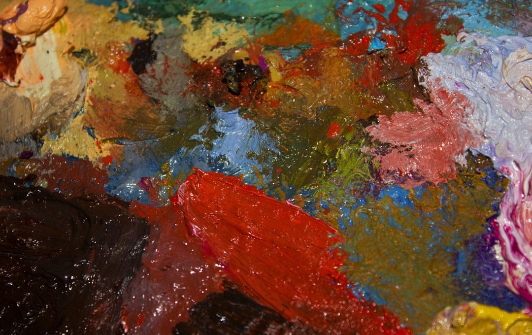 Thick impasto oil paint swirls in red, blue, and yellow with heavy texture on an artist's wooden palette.