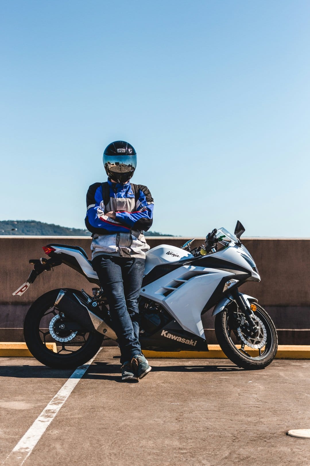 Motorcyclist in a black helmet and jacket leans against a white Kawasaki bike on concrete under a blue sky.