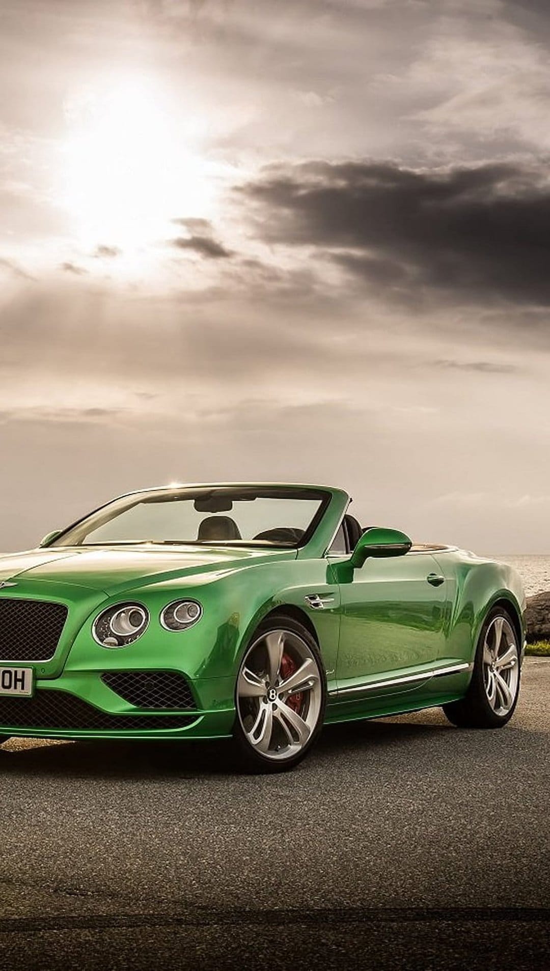Emerald green Bentley Continental GT Convertible parked on a coastal road under a dramatic cloudy sky by the sea.