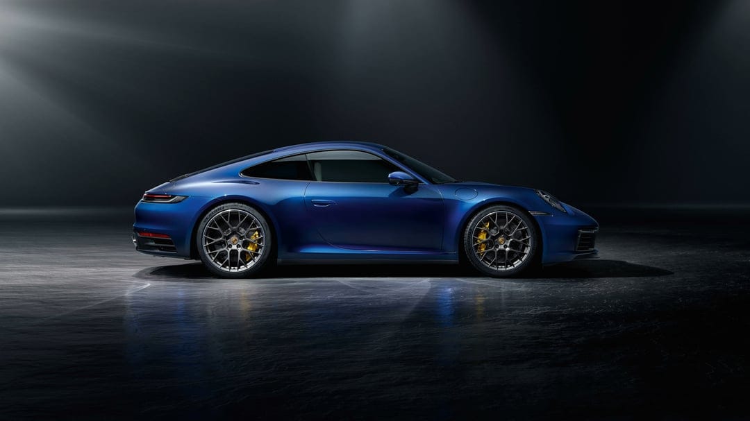 Blue Porsche 911 side profile with yellow brake calipers in a dark studio under dramatic overhead lighting.