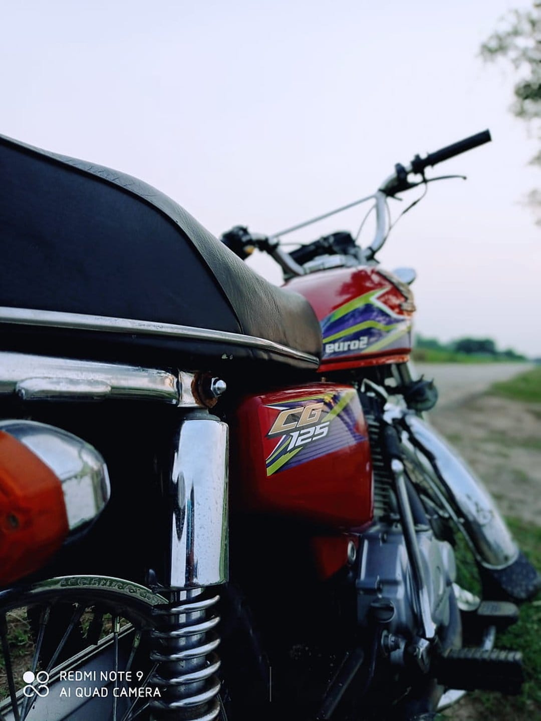 Red Honda CB 125 motorcycle parked on grass with chrome details and a dirt road background under a clear sky.