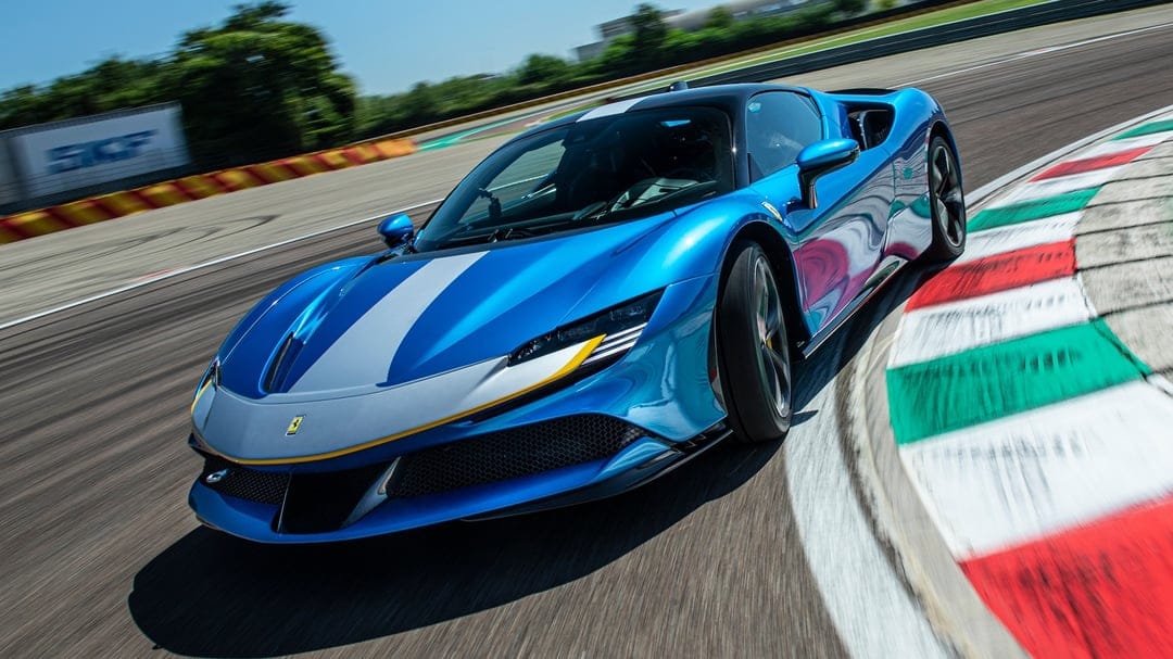 Blue Ferrari SF90 Stradale with silver racing stripes driving fast over colorful track curbs in daylight.