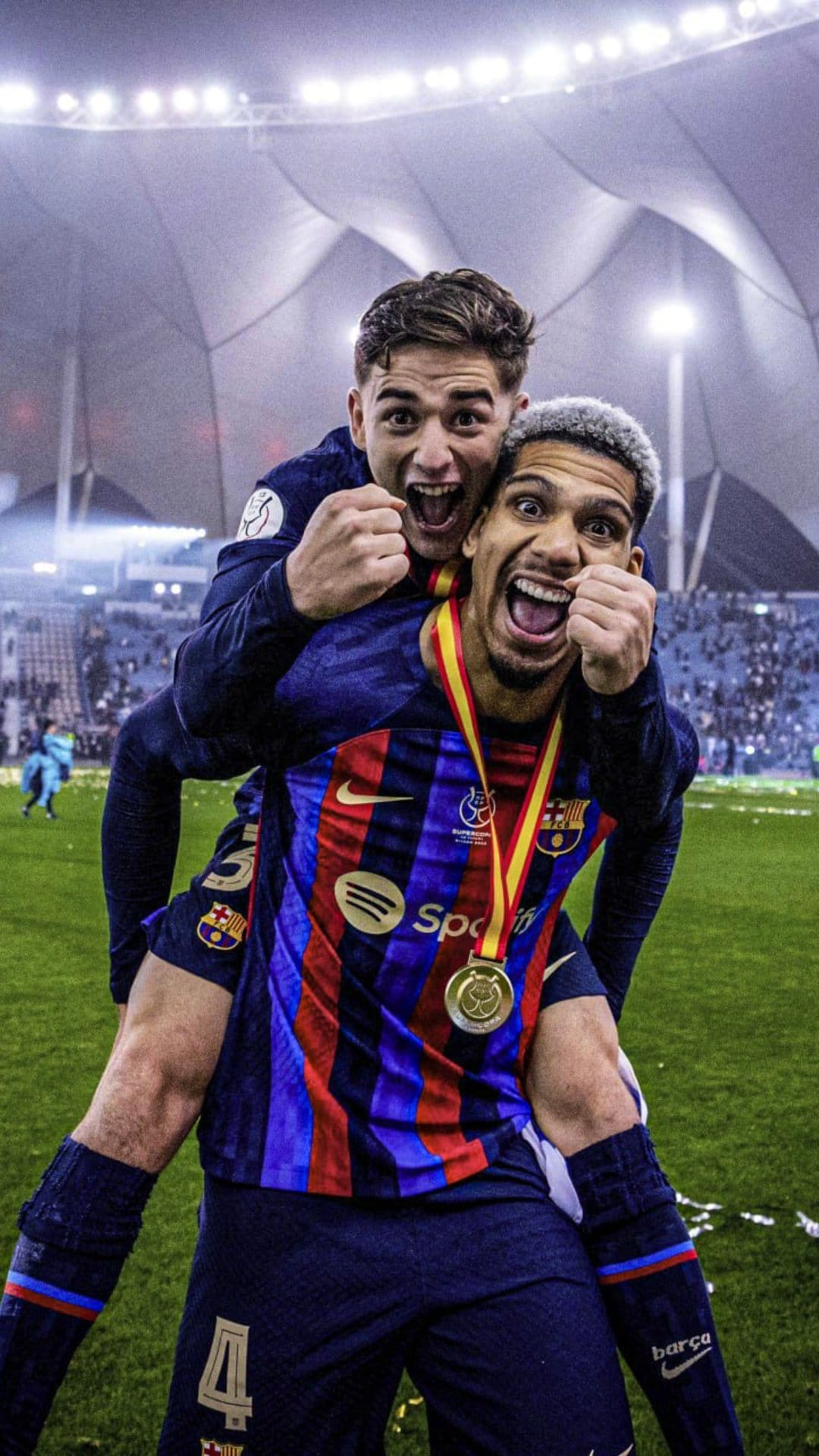 Two soccer players in blue and red jerseys hug while wearing gold medals amidst bright stadium lights at night.