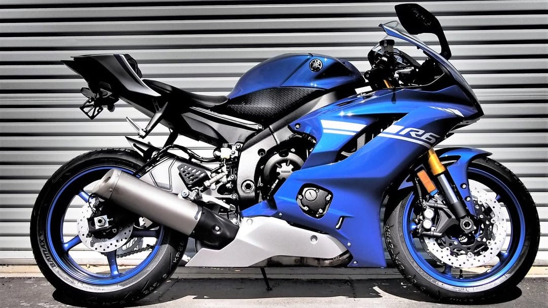 Blue Yamaha R6 sportbike side profile against a gray metal shutter with metallic accents and blue wheels.