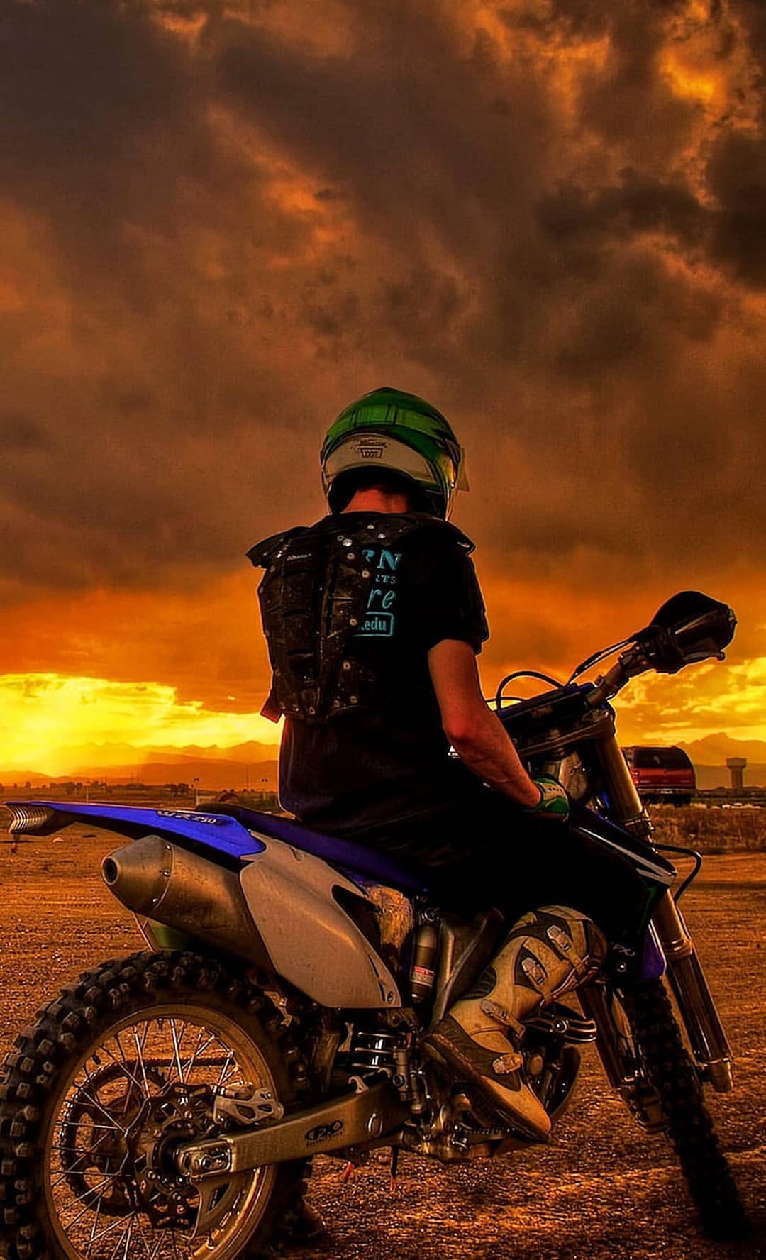 Motocross rider in a green helmet on a blue dirt bike overlooks a mountain range during a vibrant orange sunset.