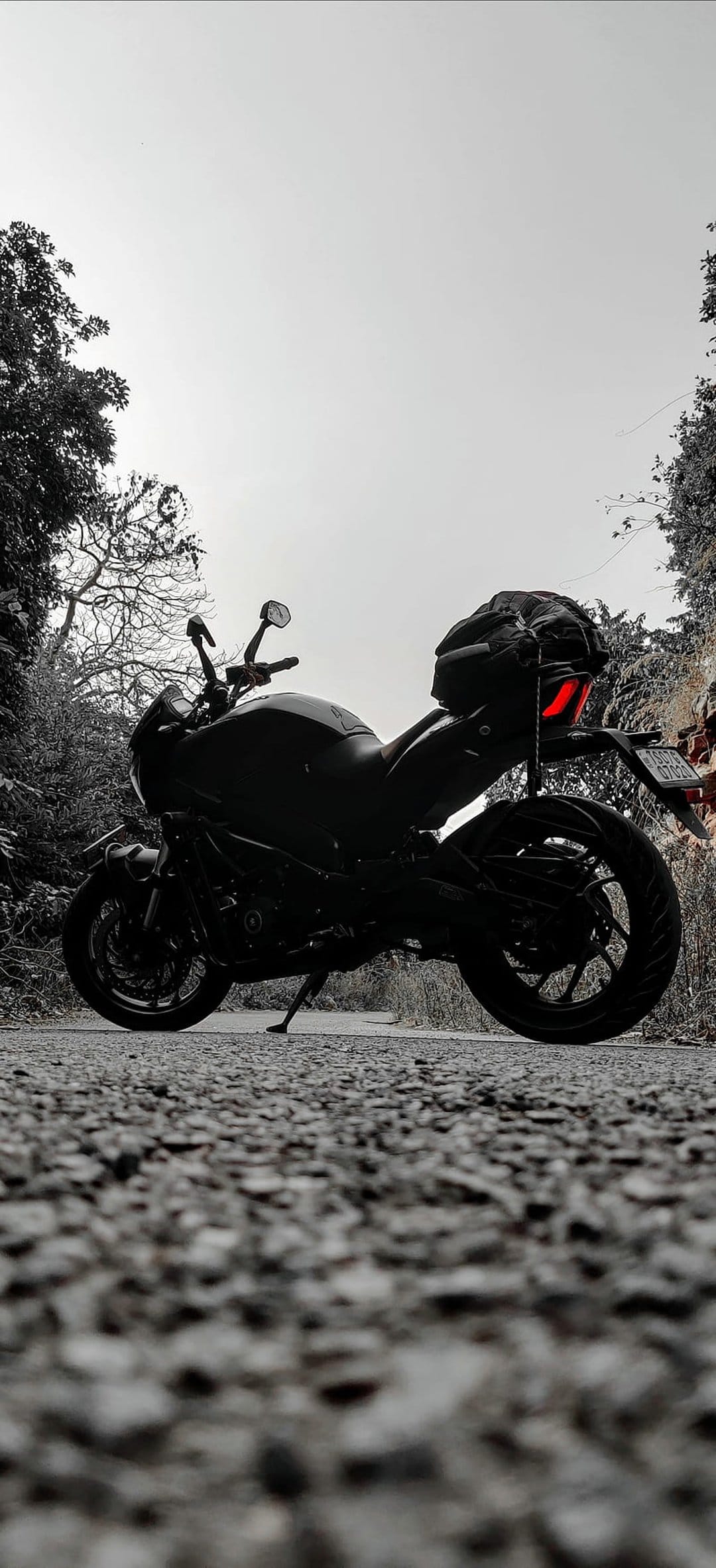 Motorcycle silhouette on a textured gravel road with a glowing red brake light and trees in the distance.