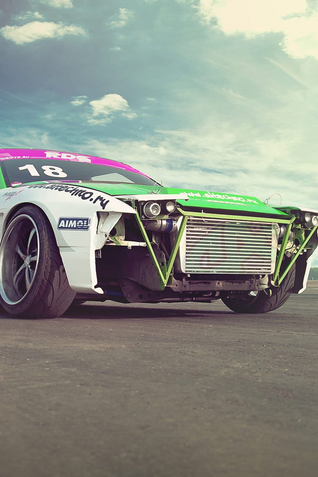 Drift car number 18 features a front intercooler, neon green roll cage, and pink livery against a cloudy sky.