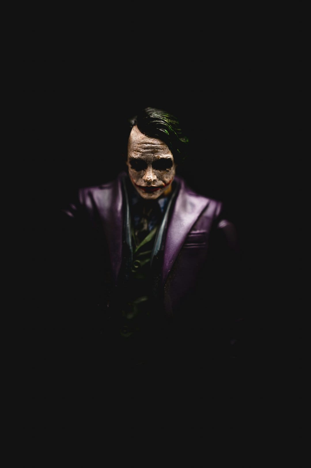 Joker figurine with green hair and purple suit stares intensely from a dark and heavily shadowed background.