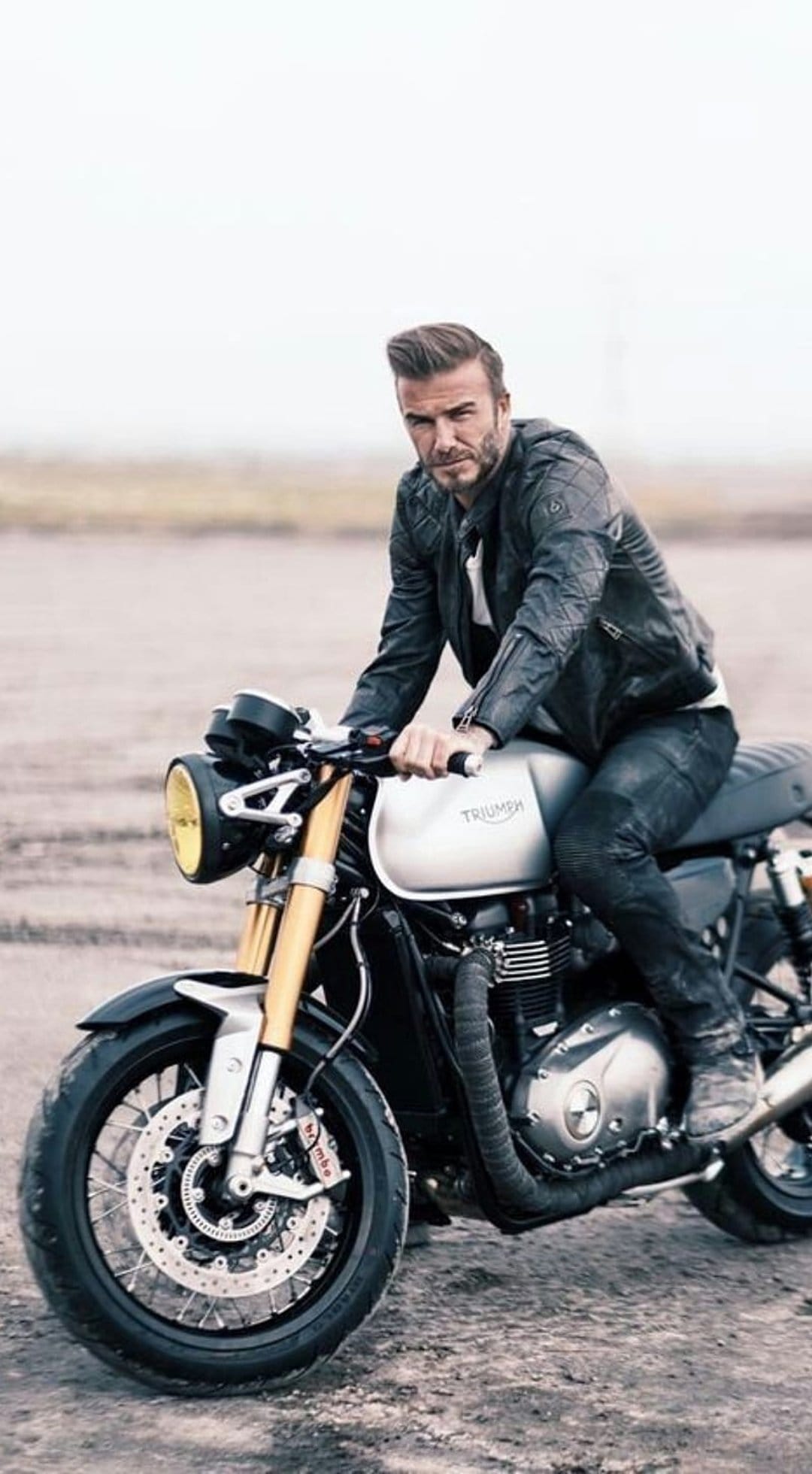 David Beckham in a leather jacket sits on a Triumph motorcycle against a blurred outdoor background in this portrait.