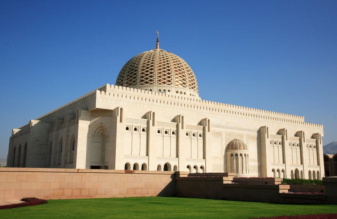 Sultan Qaboos Grand Mosque features a massive dome and tall minarets against a clear blue sky in Muscat Oman.