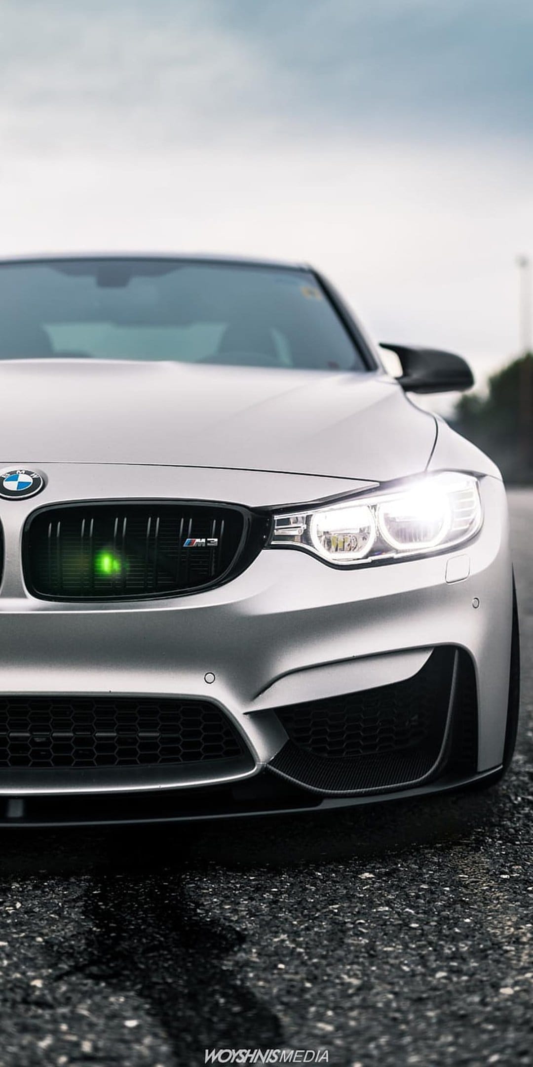Silver BMW M3 front grille and glowing LED headlights on a damp asphalt road featuring neon green accents.