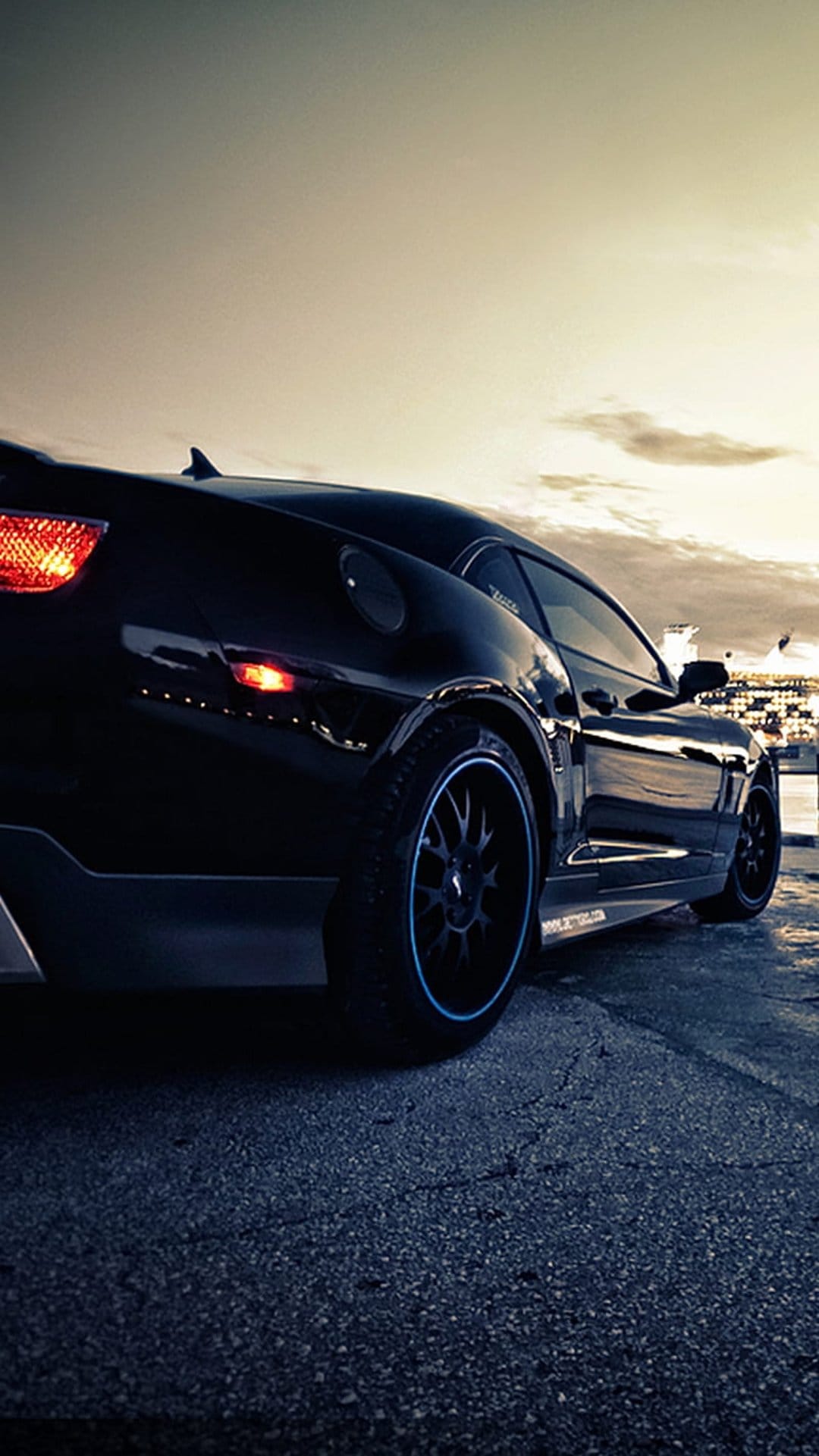 Black muscle car with illuminated red taillights sits on asphalt by water under a cloudy dusk sky at evening.