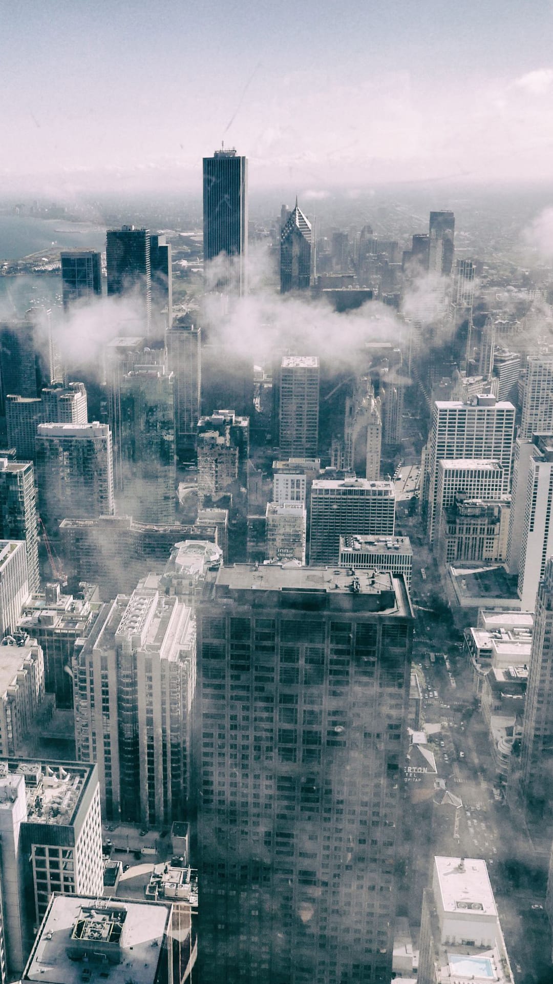 Modern skyscrapers emerge through a dense blanket of white clouds and fog under a soft, overcast light.