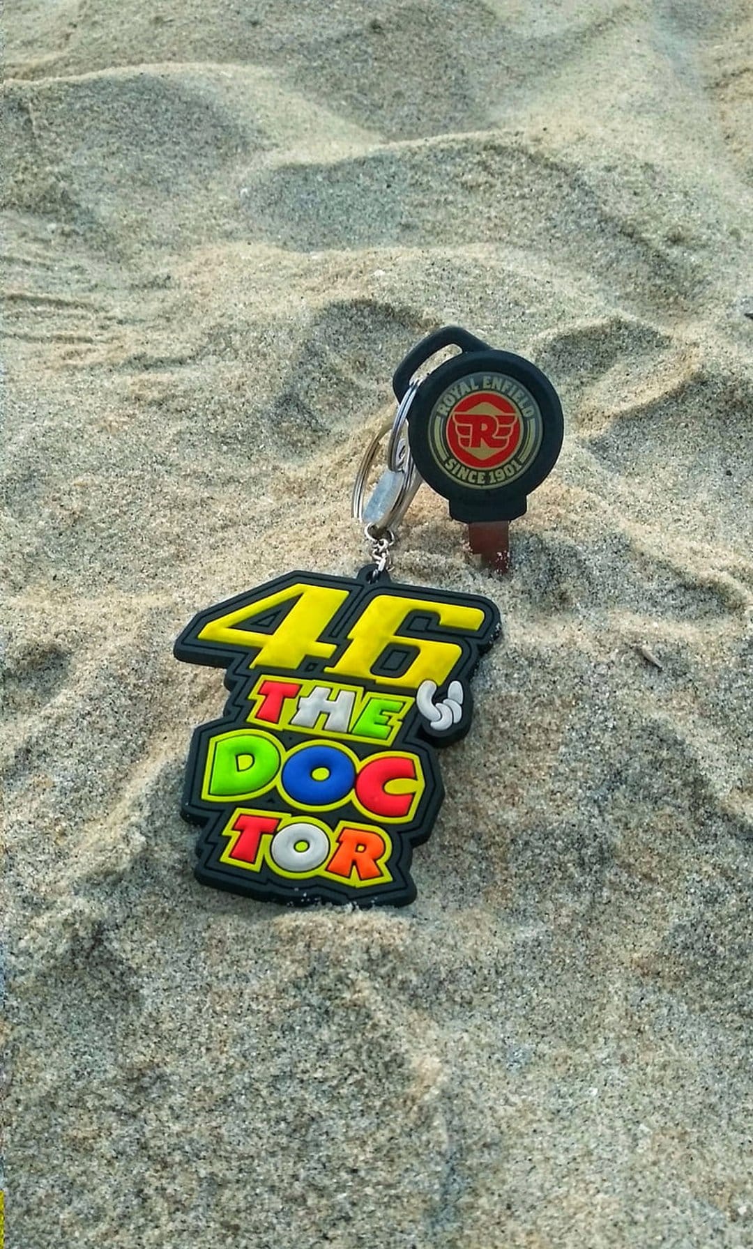 Yellow 46 keychain and Royal Enfield motorcycle key rest on fine beach sand with soft natural sunlight.