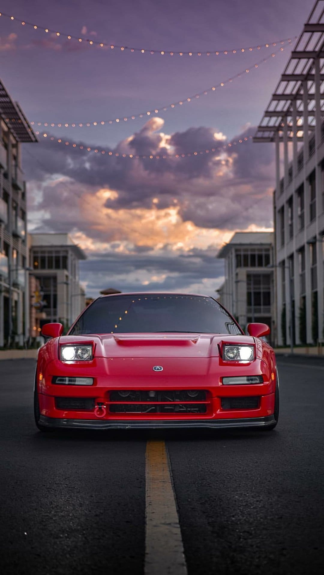 Red Acura NSX parked on a city street at dusk with string lights reflecting off the hood and a cloudy sky.