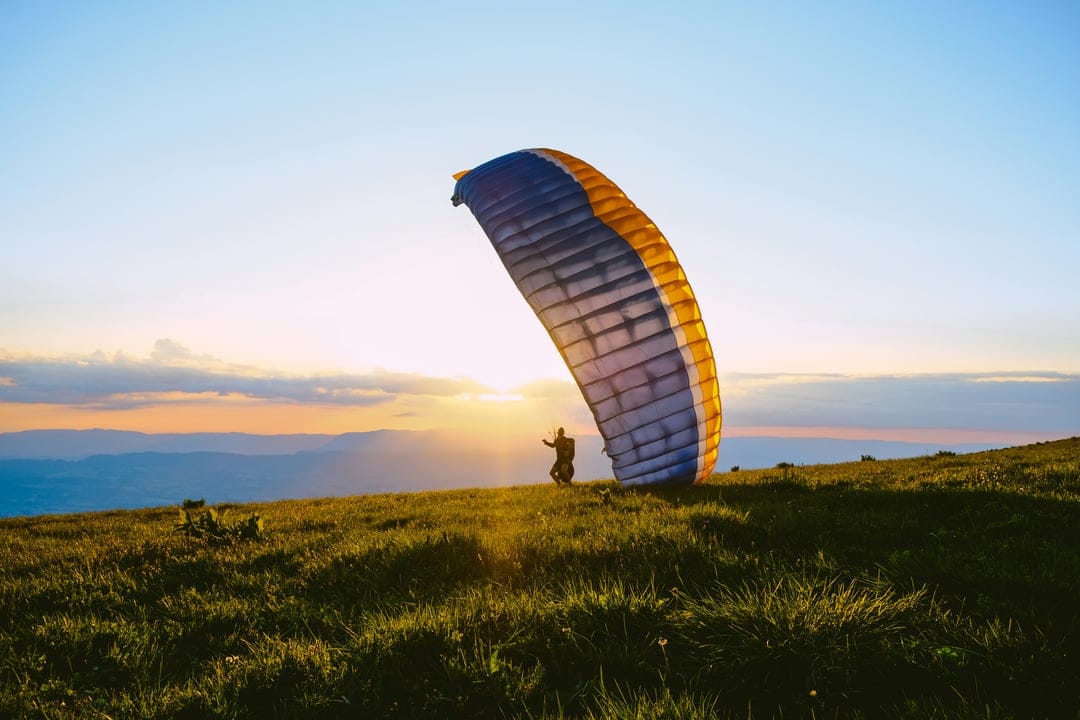 Paraglider on a grassy hill prepares to launch with an inflated wing silhouetted against a golden sunset sky.