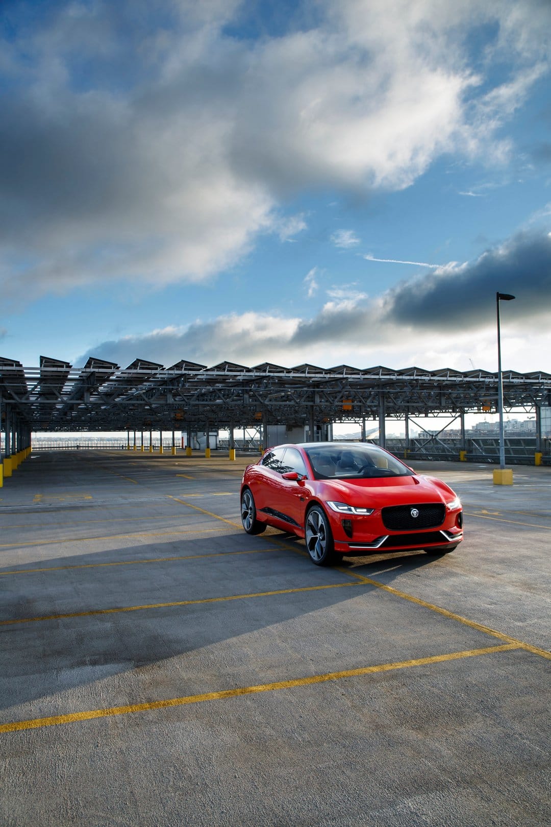 Red Jaguar I-PACE electric SUV parked in a concrete lot with solar panels under a blue, cloudy sky at an angle.