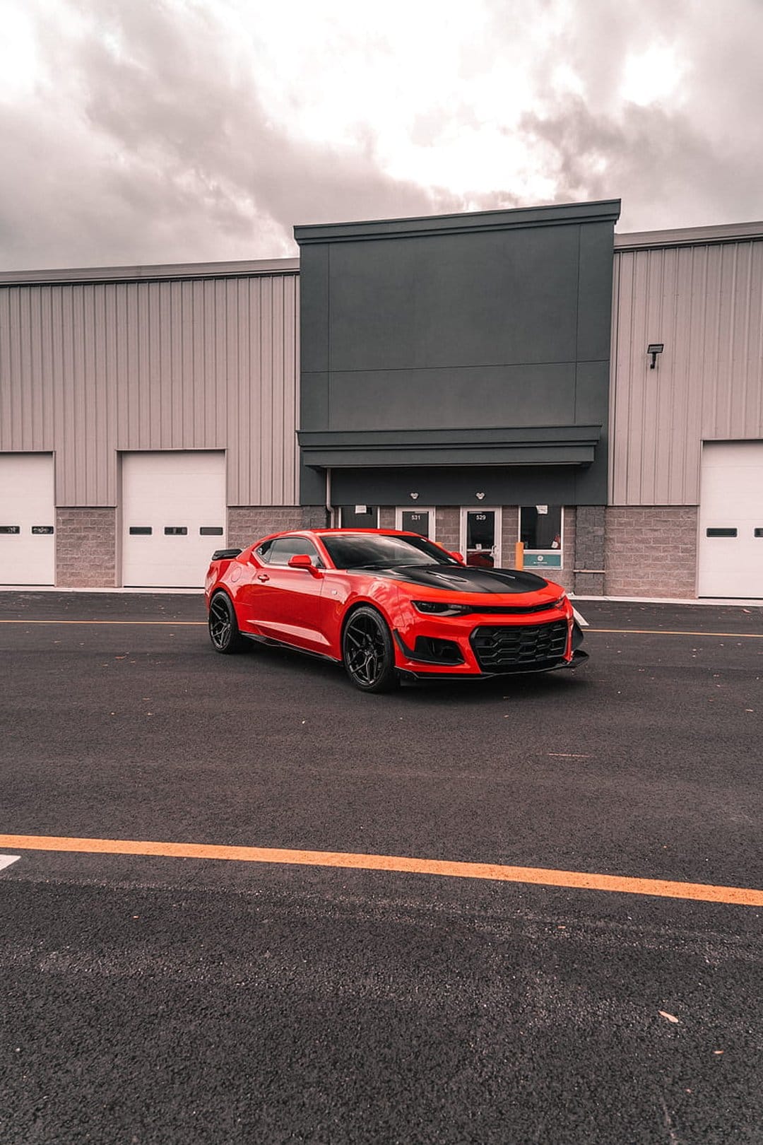 Red Chevrolet Camaro ZL1 with a black hood parked in front of white garage doors and a modern building.