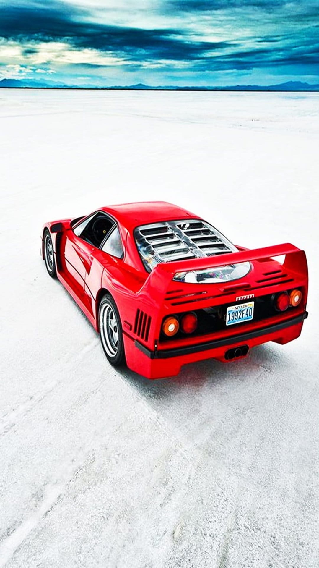 Red Ferrari F40 rear view on vast white salt flats with a dramatic cloudy sky and distant mountain peaks.