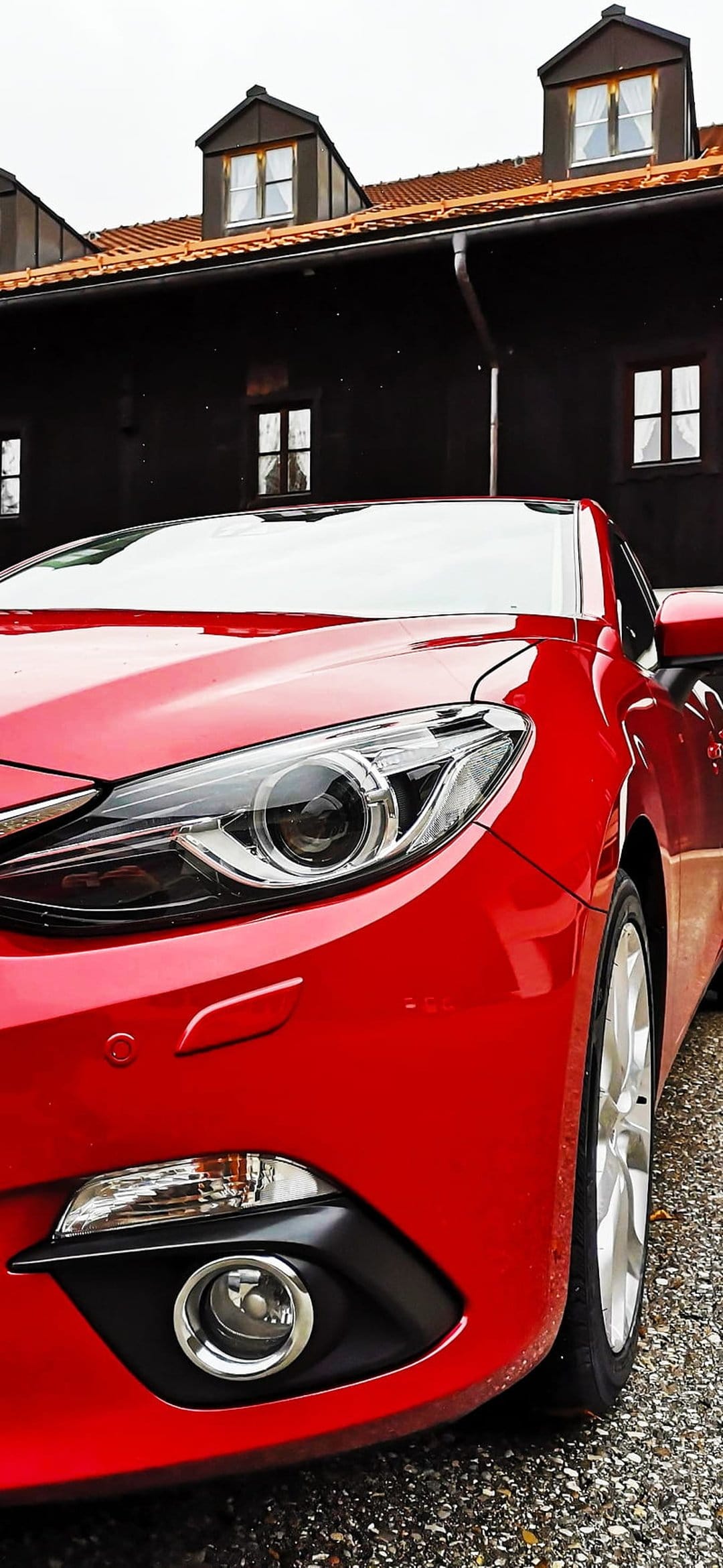Red Mazda 3 parked on gravel with a glowing headlight in front of a traditional building with a tiled roof.
