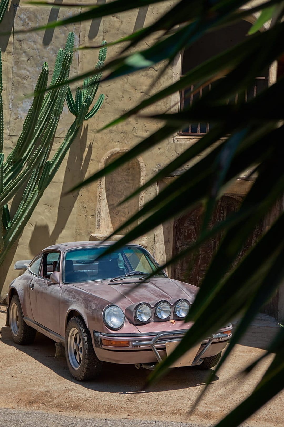 Vintage Porsche 911 rally car with off-road tires parked by a stone building and cacti in a desert oasis.