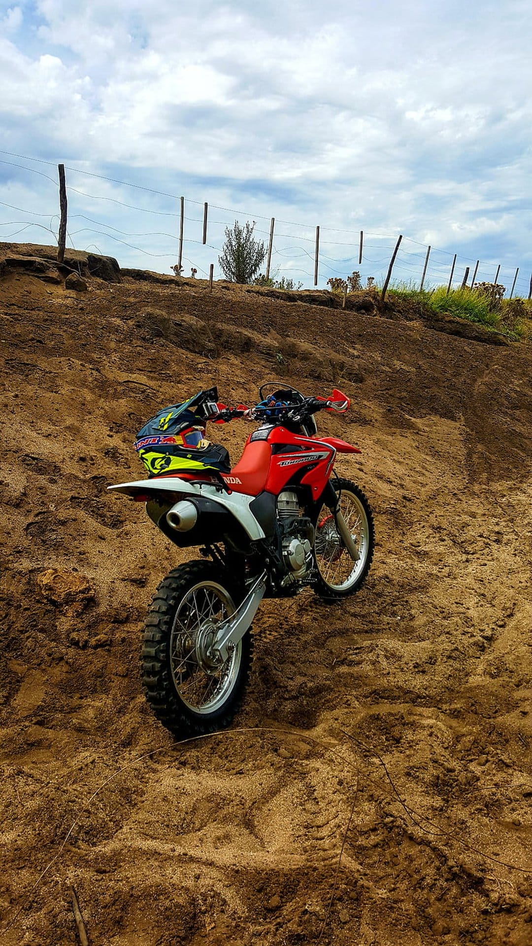 Red dirt bike with a colorful helmet parked on a sandy hill under a cloudy, gray sky in an outdoor setting.