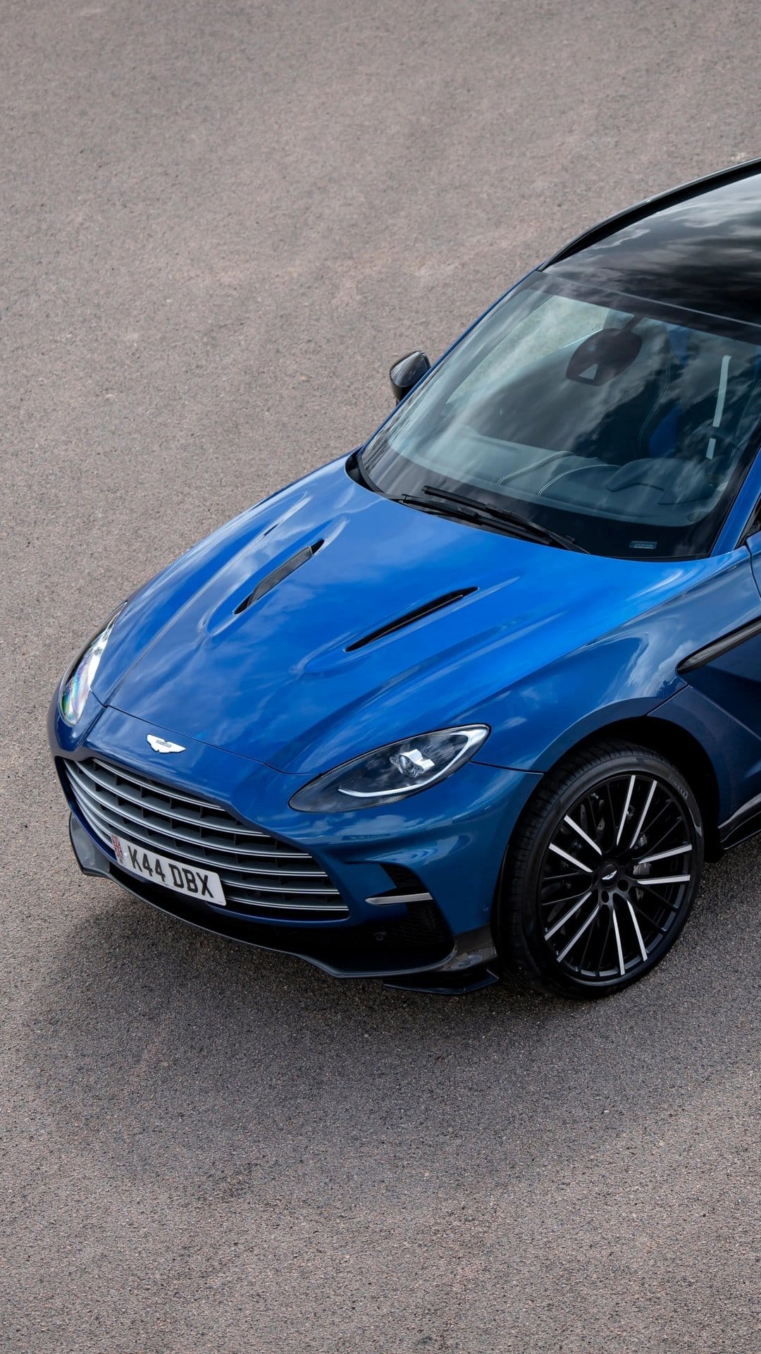 Front view of a metallic blue Aston Martin DBX on asphalt with detailed headlights and multi-spoke wheels.