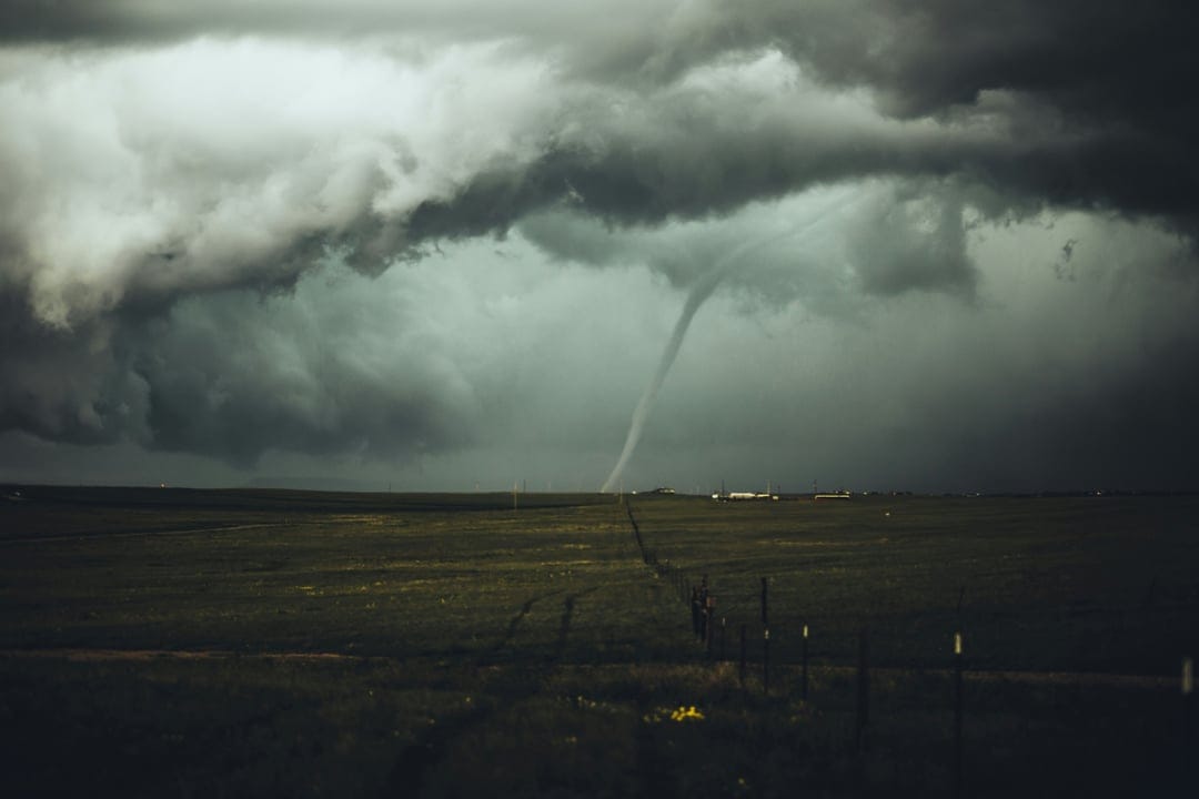 Tornado funnel cloud touching down in a flat rural field under a dark, heavy sky with a dirt road in the foreground.