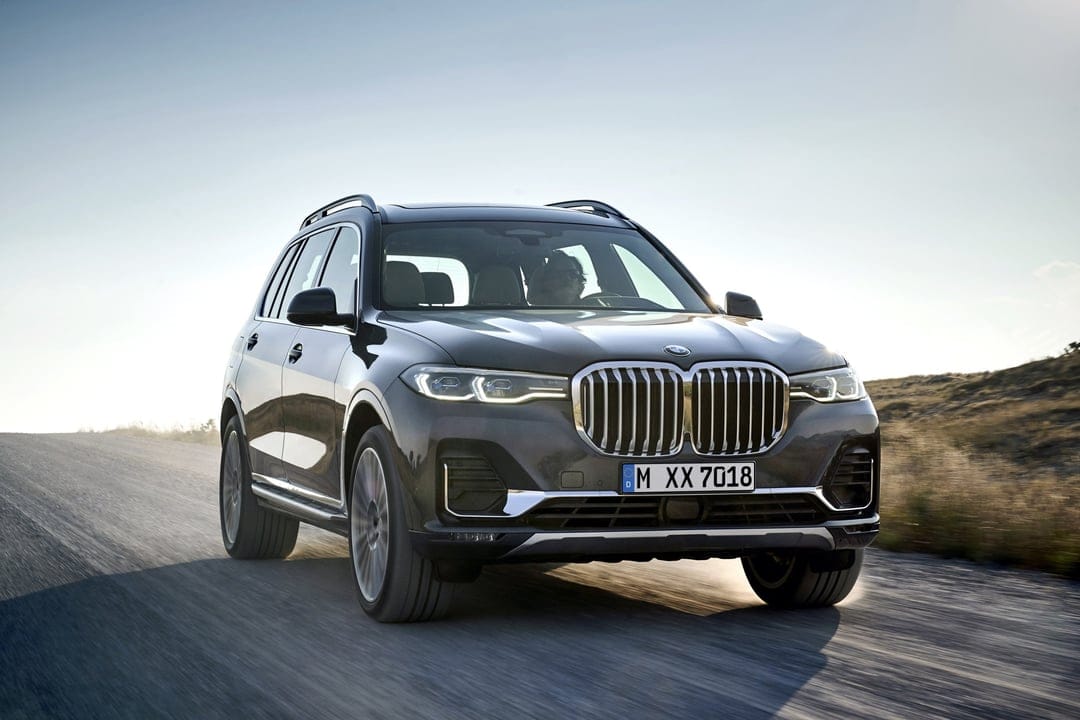 Dark grey BMW X7 SUV driving on a dusty rural road under a clear blue sky with warm sunlight highlights.