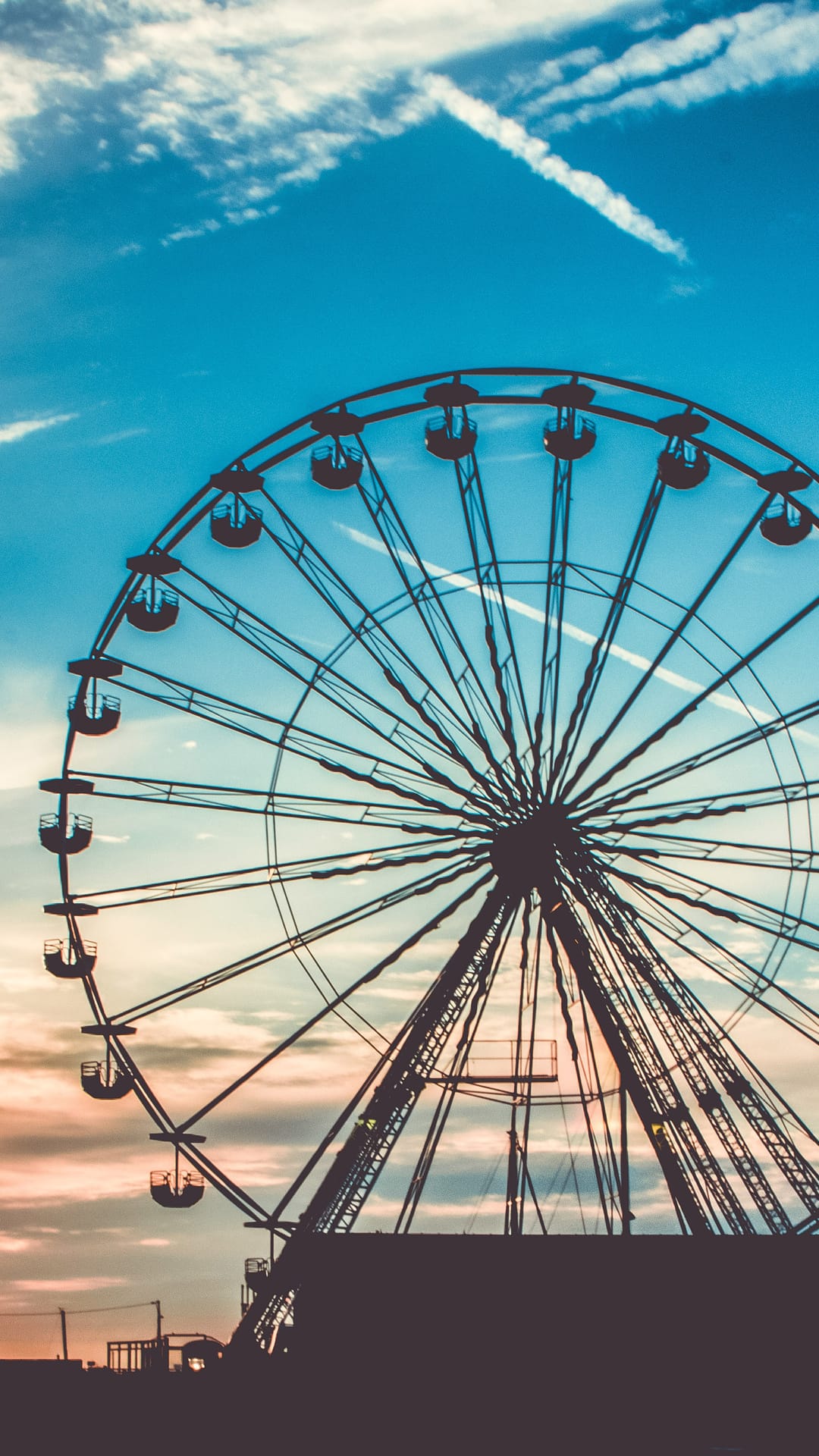 Ferris wheel silhouette against a blue and orange gradient sky featuring thin white clouds and contrails.