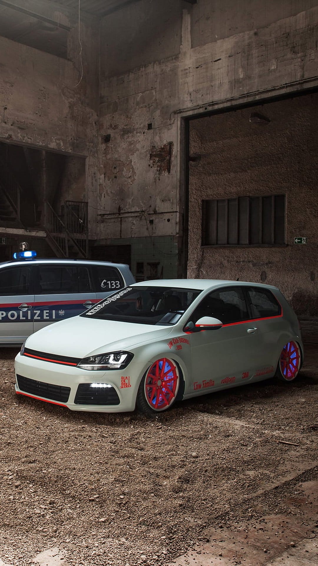 White modified VW Golf GTI with red and blue rims parked next to a police car in a gritty abandoned factory.