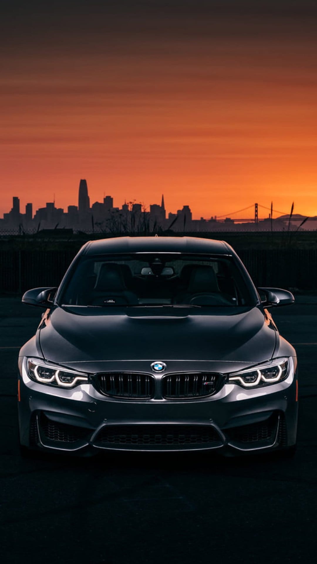 BMW M4 with glowing headlights parked before the San Francisco skyline and Golden Gate Bridge at sunset.