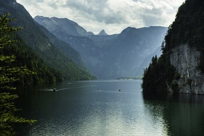 Still blue lake reflecting jagged mountain peaks and dense evergreen forests under a soft overcast sky with boats
