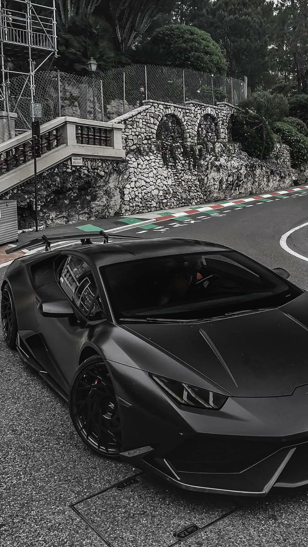 Matte black Lamborghini parked on a cobblestone Monaco street circuit with stone walls and green foliage.