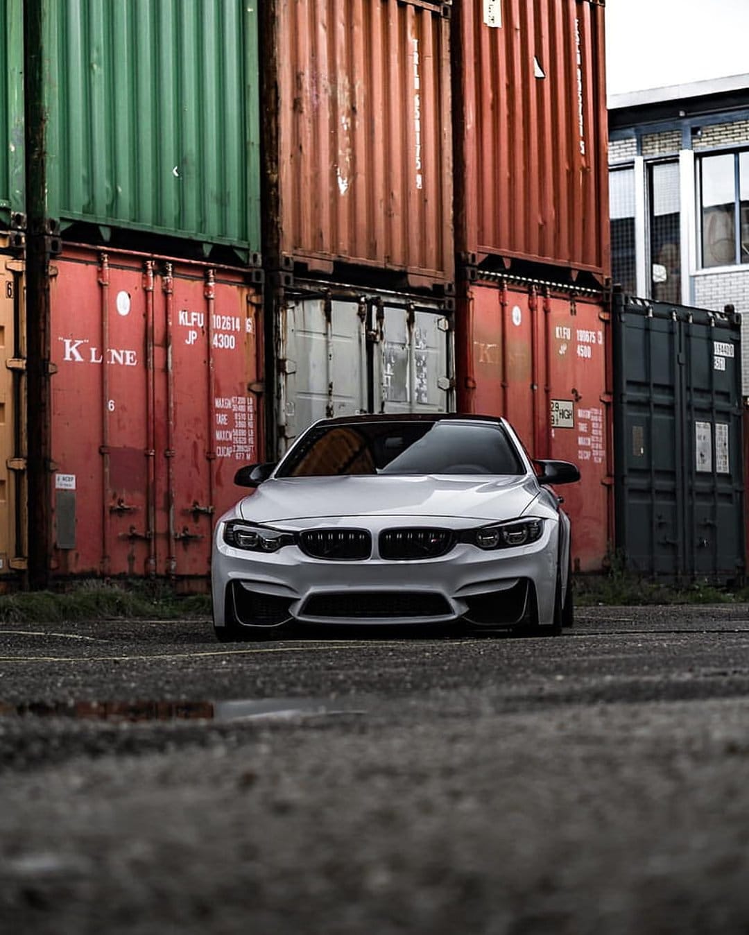 White BMW M4 sports car parked in an industrial shipping yard with colorful stacked cargo containers behind it.