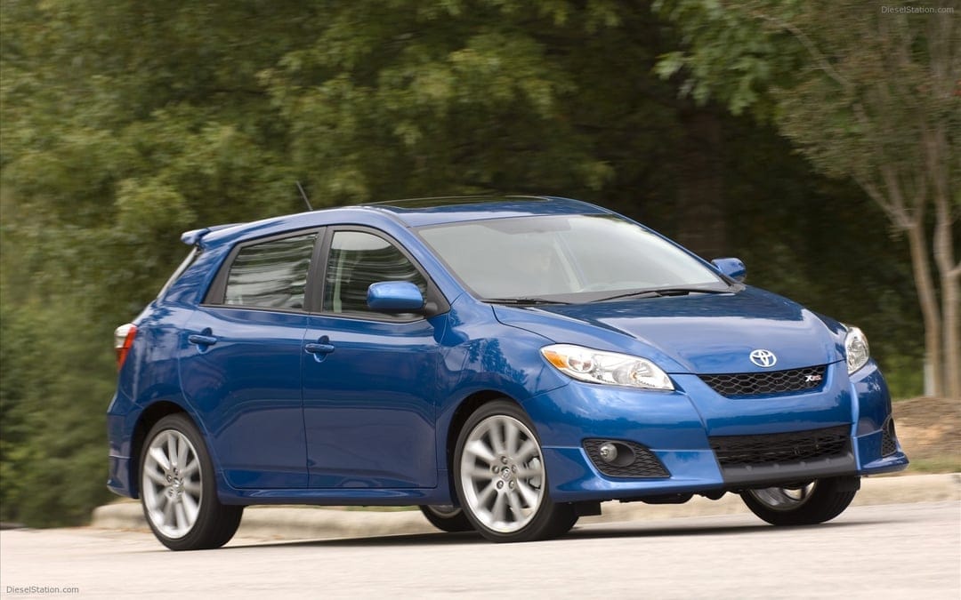 Blue Toyota Matrix XRS hatchback driving on a road with motion blurred wheels and green tree background.