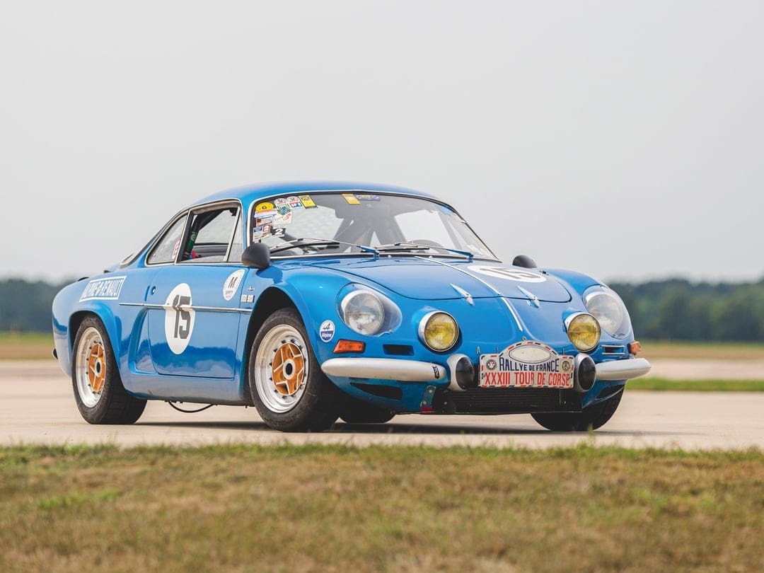Blue Alpine A110 rally car with number 15 and yellow lights parked on a wide asphalt track under a grey sky.