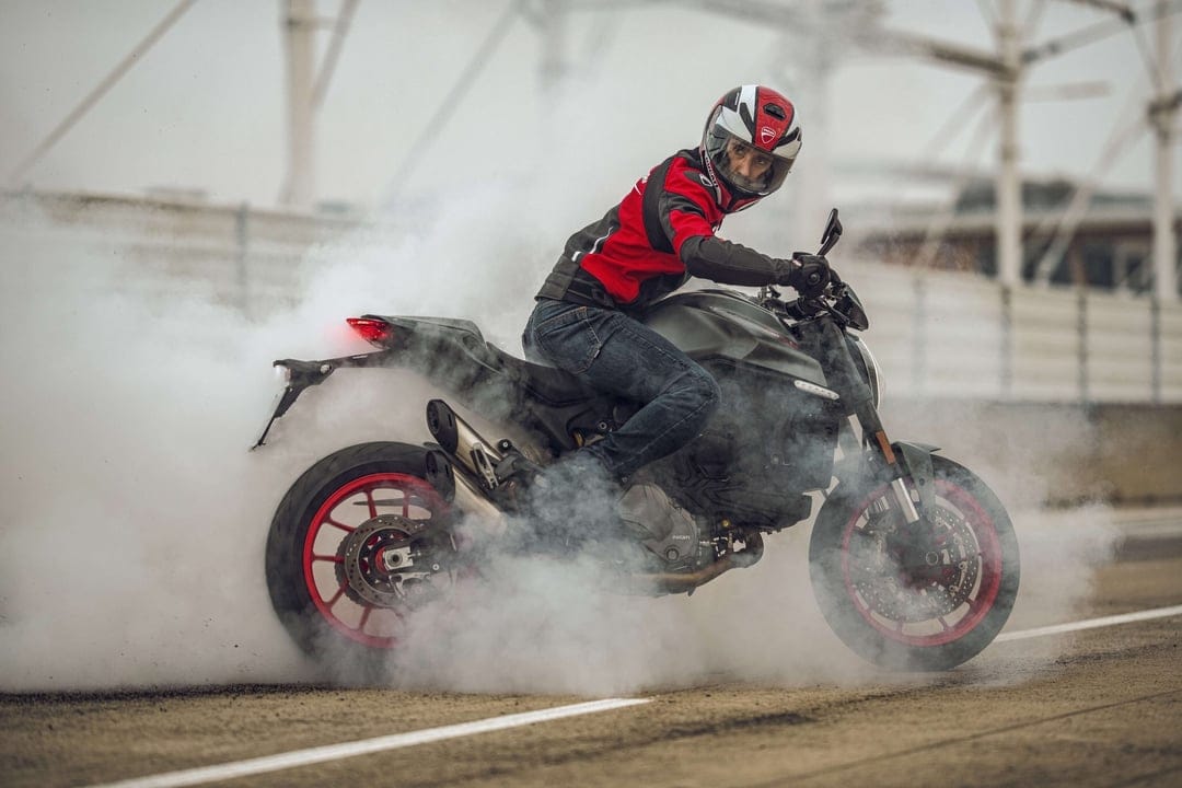 A motorcyclist on a Ducati performing a burnout with thick white smoke surrounding the red rear wheel.
