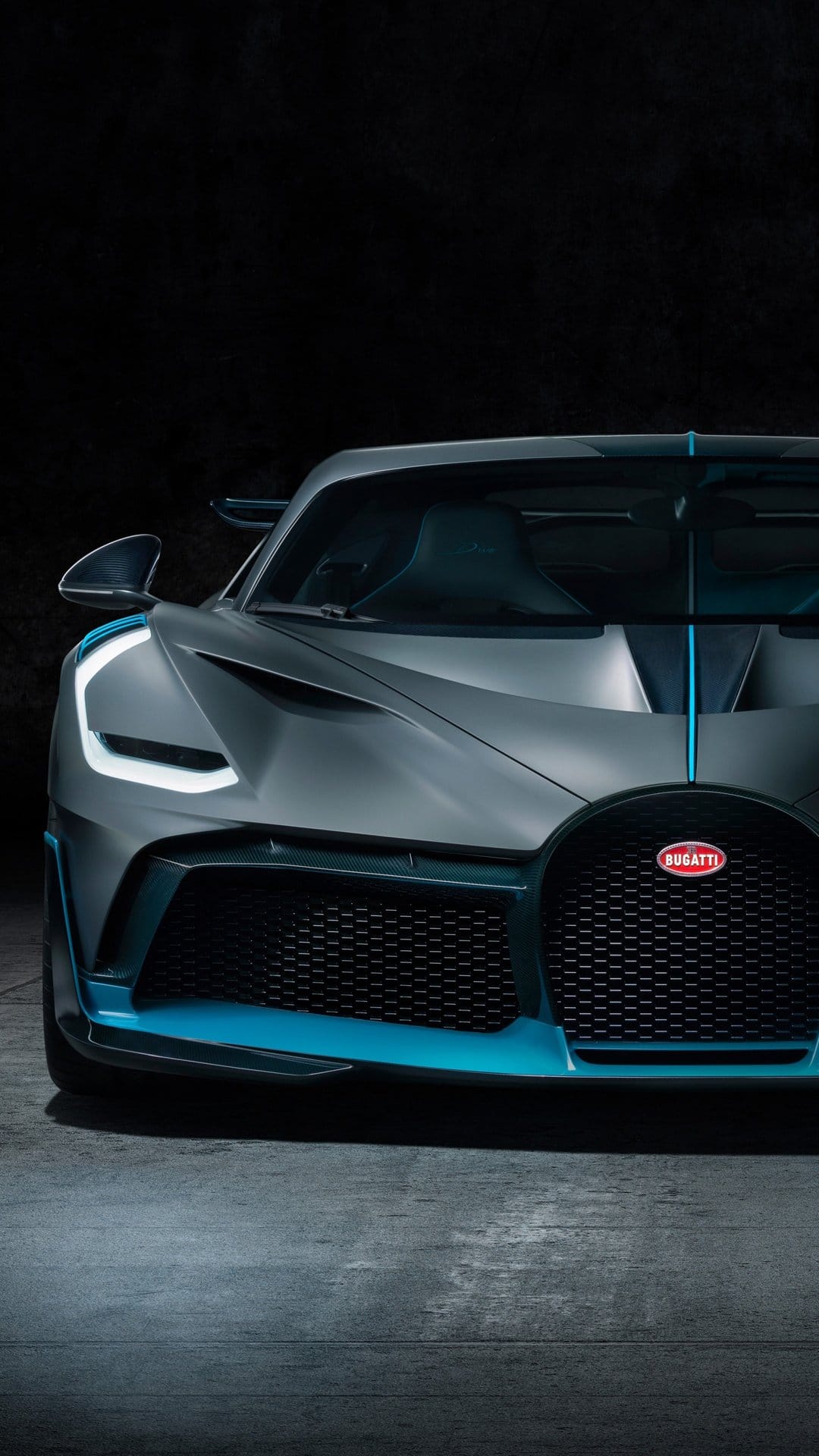 Close-up of a matte grey Bugatti Divo with turquoise trim and glowing LED headlights in a dark studio.