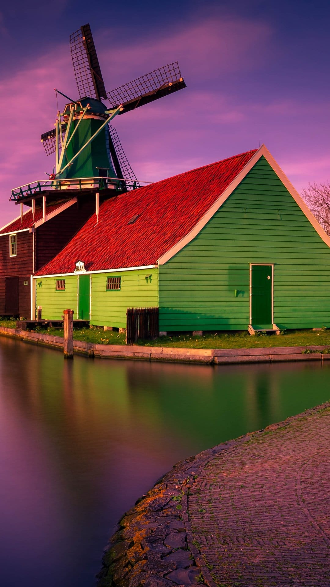 Traditional green windmill and wooden houses reflect in a still canal during a purple and orange sunset.