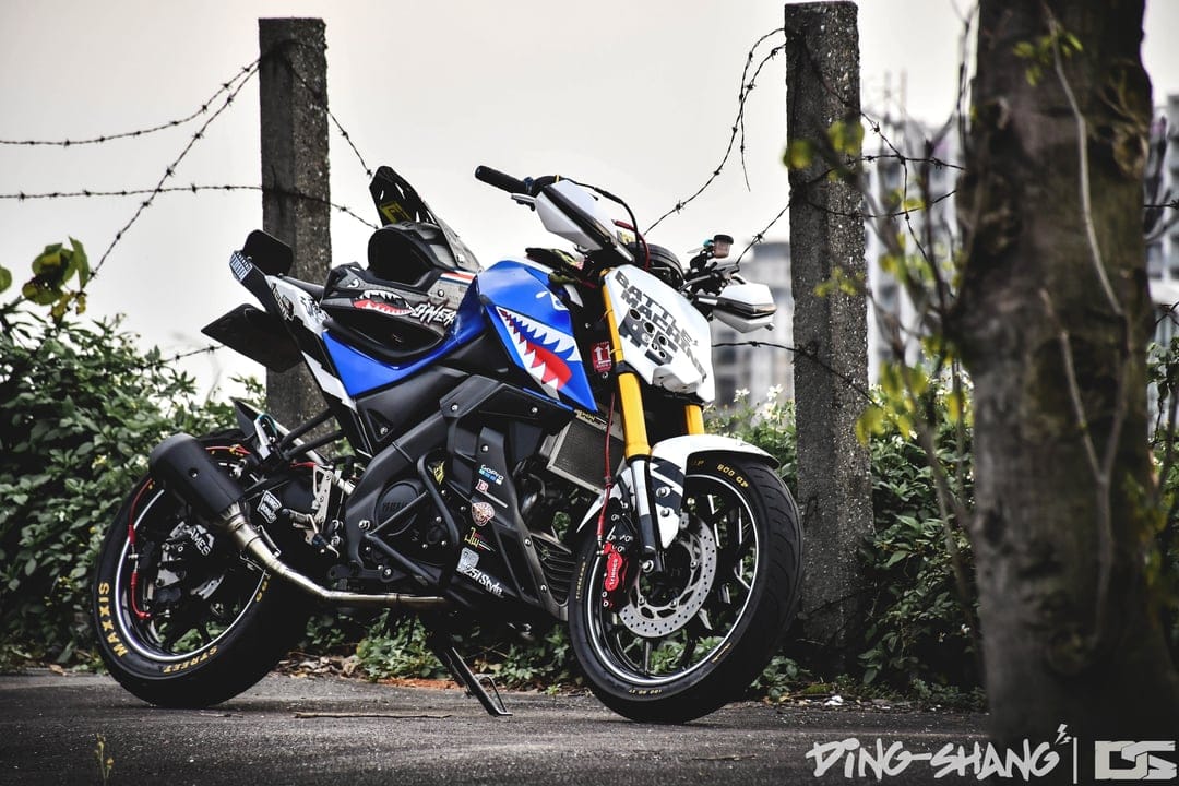 Yamaha motorcycle with blue and white shark teeth graphics and yellow suspension near a barbed wire fence.
