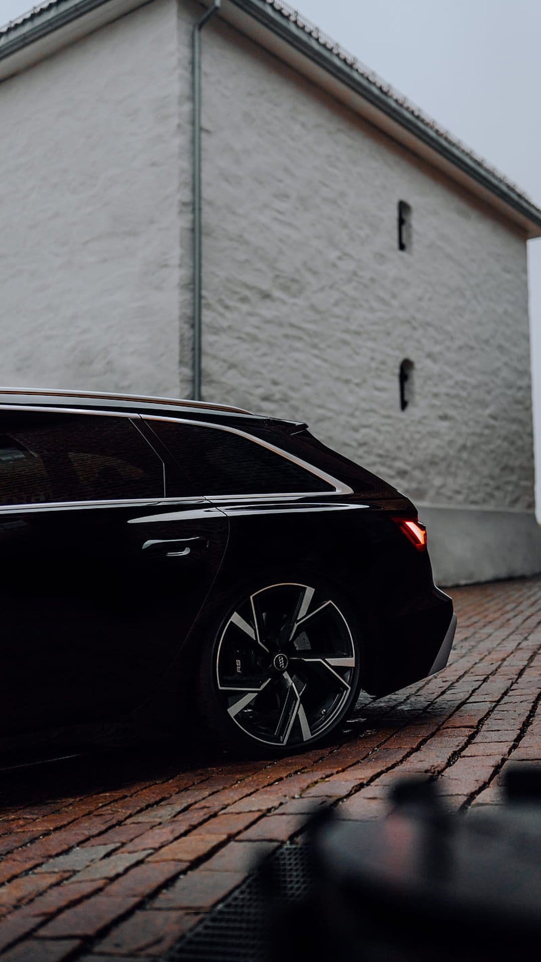 Black Audi RS6 Avant station wagon parked on rain-slicked brick street next to a white textured building.