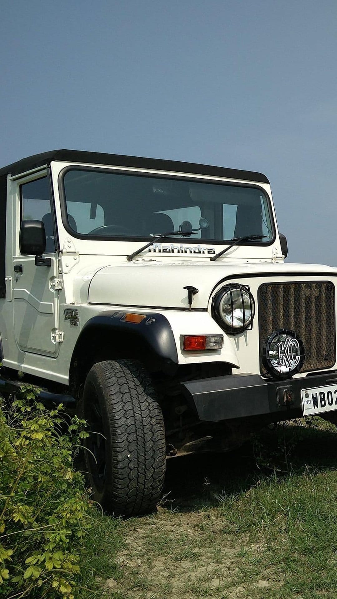 White Mahindra Thar 4x4 SUV parked in a verdant field under a bright blue sky with off-road tires visible.
