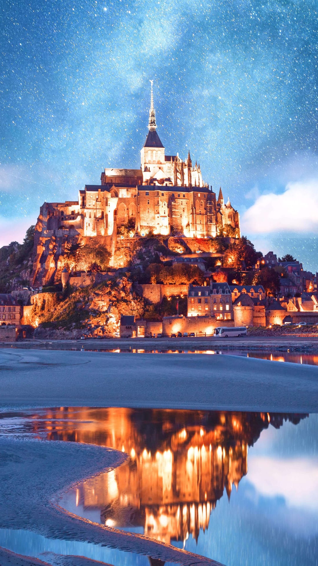 Mont Saint-Michel abbey glows with warm lights under a starry night sky with water reflections in the bay below.