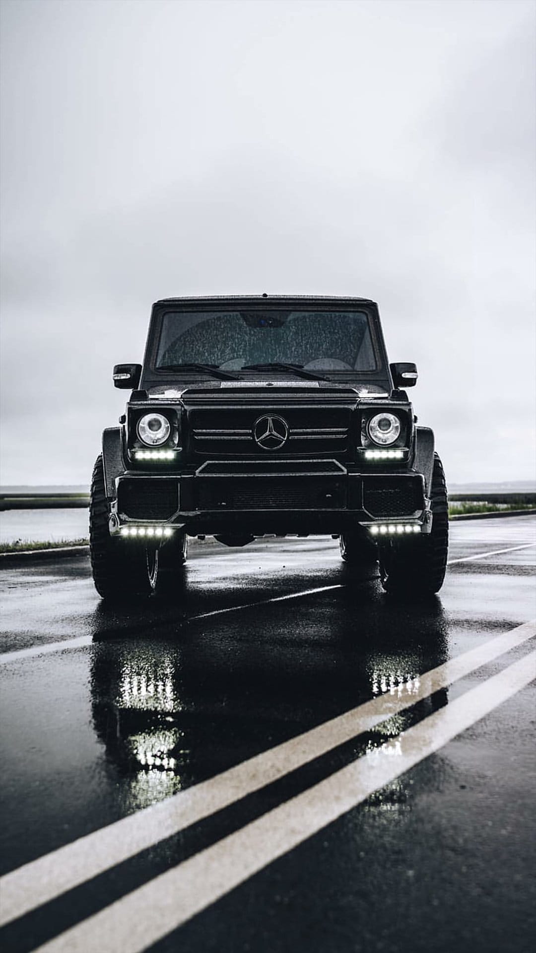 Black Mercedes G-Wagon parked on a wet road at dusk with illuminated headlights and reflections on the pavement.