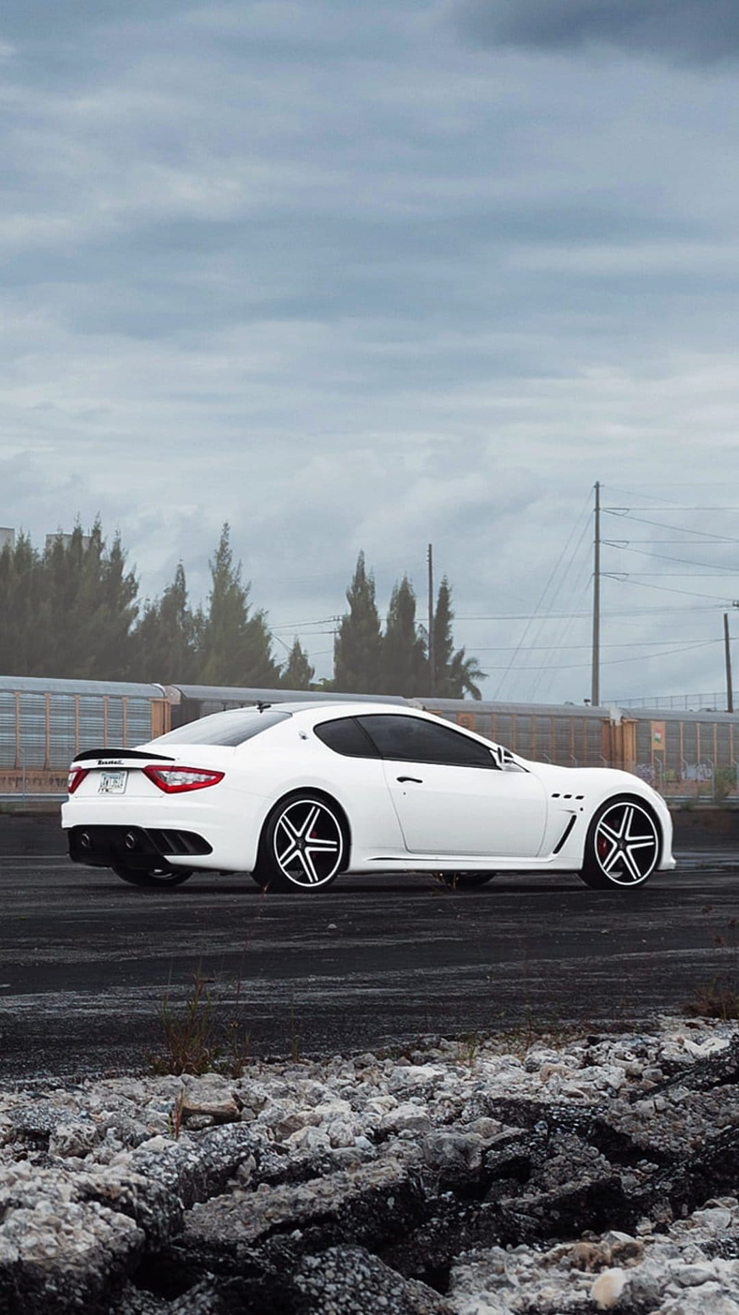 White Maserati GranTurismo side profile on asphalt with custom wheels under a dark, stormy, overcast sky.