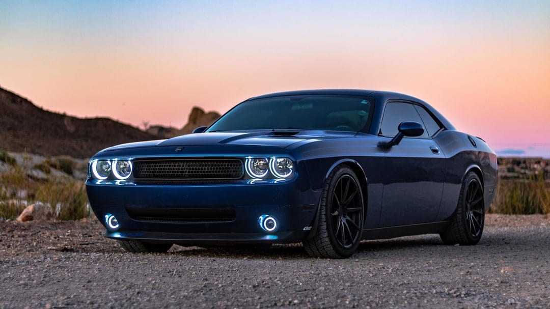 Blue Dodge Challenger with glowing halo headlights on a gravel road during a pink and orange desert sunset.