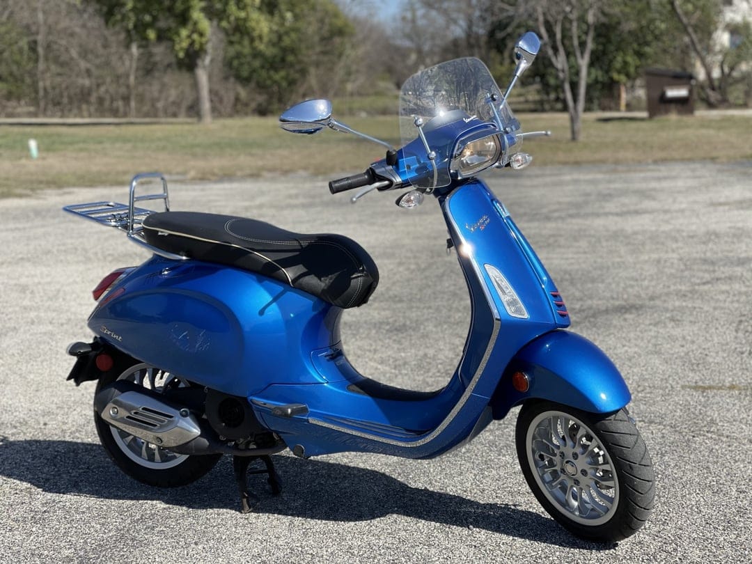 Blue Vespa Sprint scooter with chrome accents and windshield parked on asphalt with blurred trees in the background.