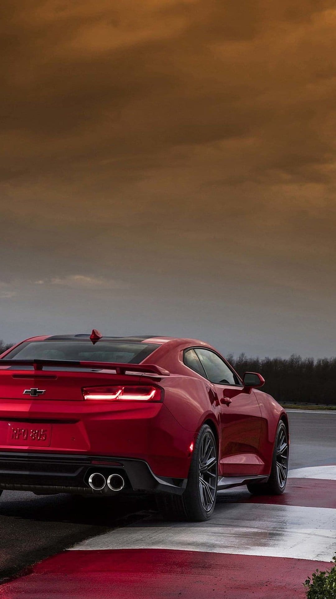 Rear of a red Chevrolet Camaro with lit brake lights on a wet racing circuit under a dramatic overcast sky.