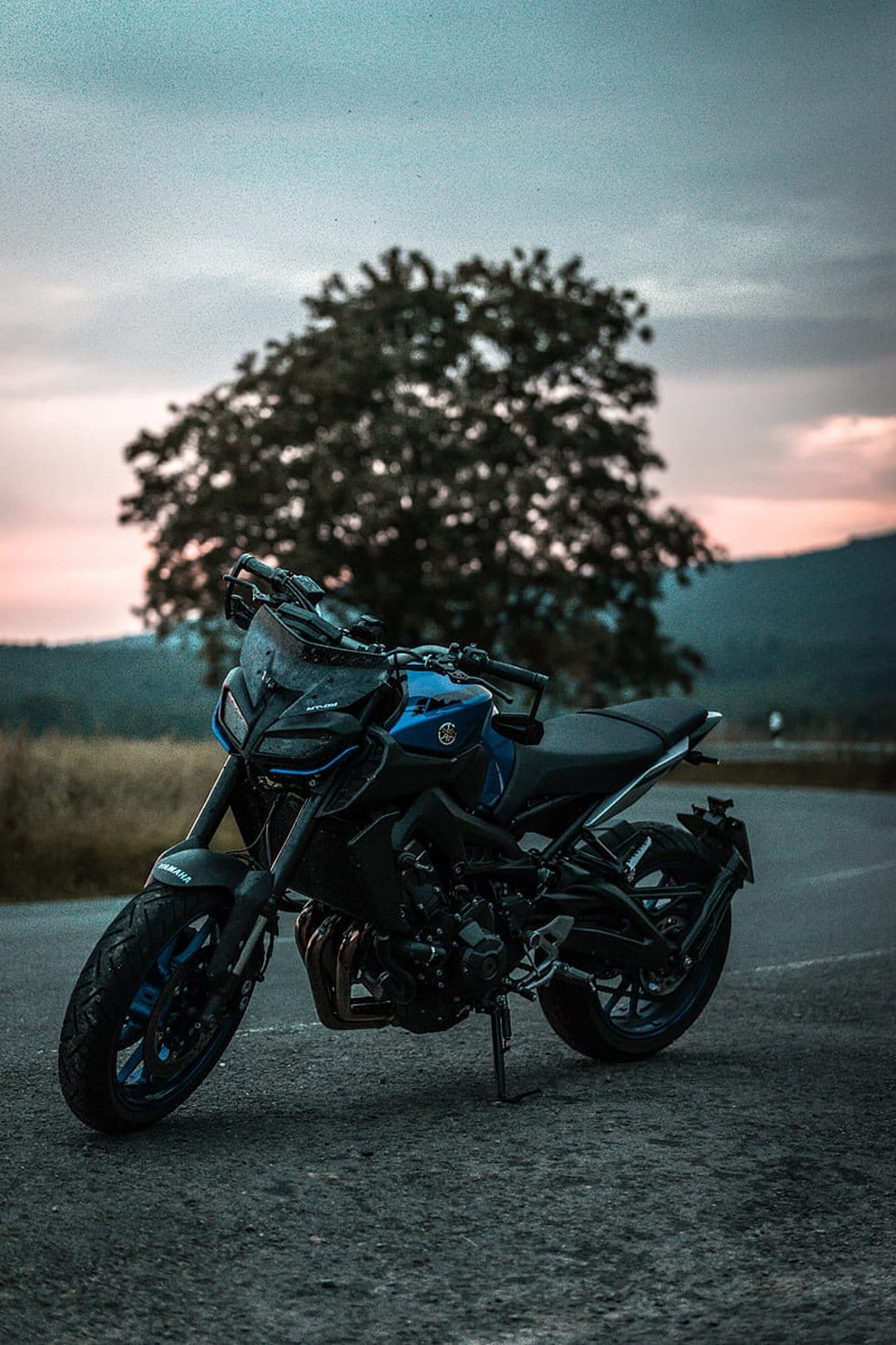 Blue Yamaha MT-09 motorcycle parked on a paved rural road with a silhouetted tree against a pink and gray dusk sky.